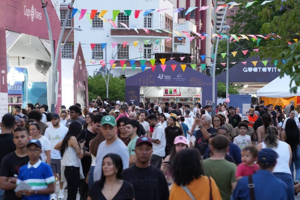 Vista panorámica de una multitud de visitantes caminando por un paseo al aire libre decorado con guirnaldas de banderines de colores, flanqueado por stands de editoriales como Grupo Planeta, Urano y Guatavita durante una feria del libro en un entorno urbano.