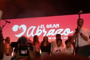 Un grupo de personas, incluidos niños y adultos, en el escenario del evento "El Gran Abrazo por Cali y el Valle". Al fondo se observa una gran pantalla LED roja con el logotipo del evento que muestra un corazón con una mano y las palabras "EL GRAN Abrazo POR CALI Y EL VALLE". Un hombre habla por un micrófono a la derecha, mientras la multitud y los equipos de grabación los rodean.